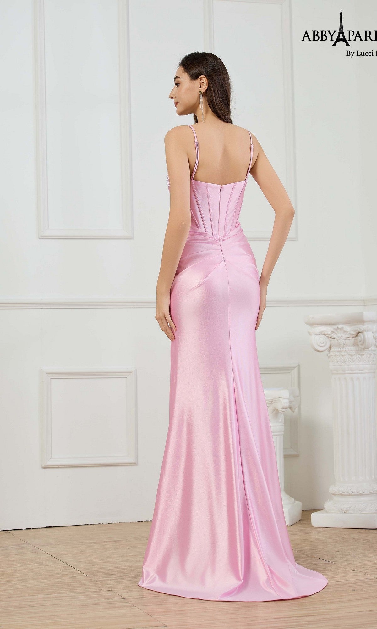 A woman stands indoors seen from the back, wearing the Abby Paris 90377, a fitted, floor-length light pink stretch satin prom dress with thin straps and a flattering silhouette.